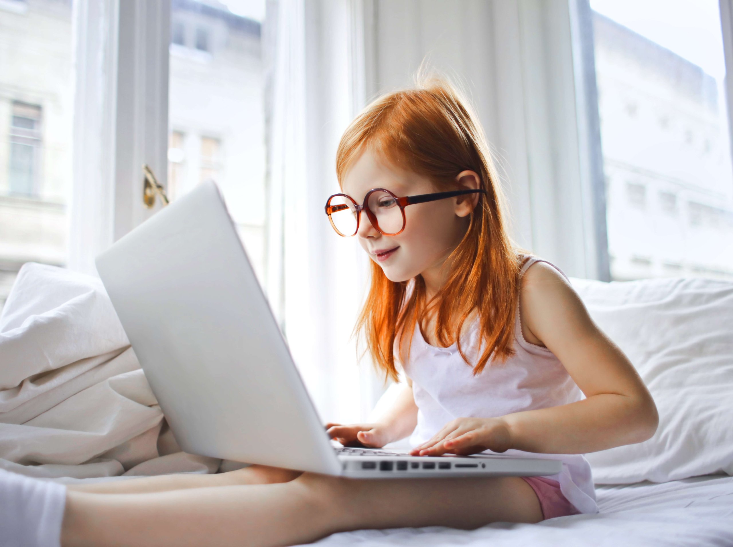 Niña con gafas sentada en la cama de su habitación jugando con el ordenador portátil de su madre